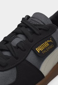 Κοντινό πλάνο μαύρου και γκρι παπουτσιού Puma Palermo με μαύρα κορδόνια και χρυσό λογότυπο Puma στο πλάι.