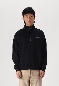 Sweatshirt noir avec un col haut et une demi-fermeture éclair, présentant un petit logo blanc sur la poitrine gauche. Fabriqué à partir d'un tissu doux et texturé.