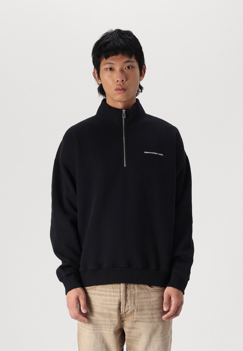 Sweatshirt noir avec un col haut et une demi-fermeture éclair, présentant un petit logo blanc sur la poitrine gauche. Fabriqué à partir d'un tissu doux et texturé.