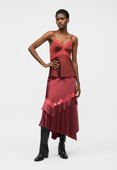Vrouw die een bordeauxrode satijnen slipjurk met kanten rand draagt en zwarte enkellaarsjes met hakken tot halverwege de kuit, staand met één hand op de heup.