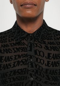 Black sheer shirt featuring a repeating "JEANS" pattern, button closures, and a classic collar, showcasing a soft, textured fabric.