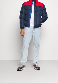 Man i marinblå och röd puffjacka, vit T-shirt, ljusblåjeans och svarta, vita och röda sneakers som står mot en enfärgad bakgrund.