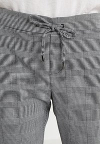 Pantalon à carreaux noir et blanc avec une taille à cordon doté d'extrémités en métal, attaché en nœud sur un fond blanc.