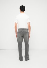 Άνδρας με πλάτη, φοράει λευκό κοντομάνικο πουκάμισο, ξεθωριασμένο γκρι τζιν και μαύρα και λευκά παπούτσια μπροστά σε απλό λευκό φόντο.