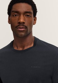 Junger schwarzer Mann mit kurzen, lockigen Haaren, der ein dunkelgraues Bugatti-Kreuzhemd trägt, vor einem schlichten Hintergrund.