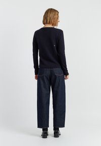 Navyblauwe gebreide sweater met een ronde hals, gecombineerd met losse, donkere spijkerbroeken. Opvallende details zijn ribgebreide manchetten en een klein logo op de achterkant.