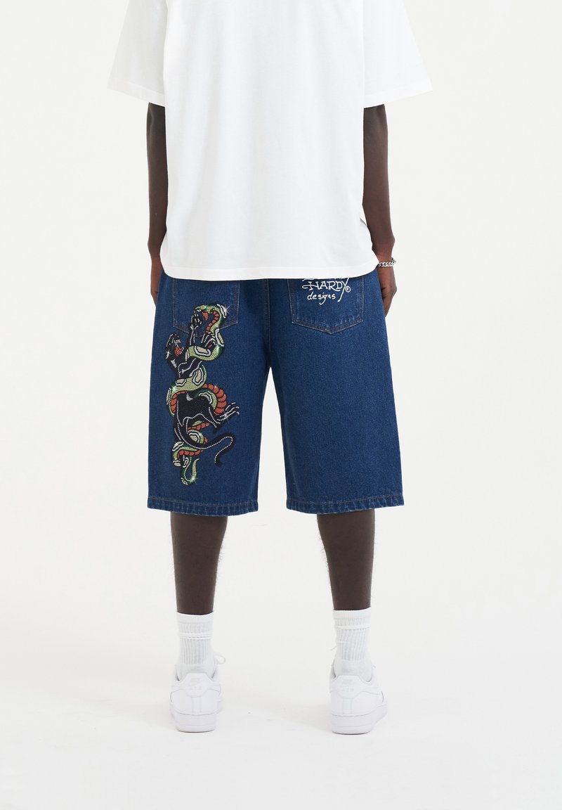 Shorts de mezclilla en azul con coloridos bordados de serpiente y pantera en la pierna izquierda, con un pequeño logo en el bolsillo trasero.