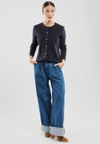 Granatowy kardigan z złotymi guzikami, zestawiony z luźnymi niebieskimi dżinsami podwiniętymi na mankietach oraz brązowymi botkami za kostkę. Gładka tekstura na całej powierzchni.