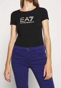 T-shirt noir à manches courtes avec logo doré EA7 Emporio Armani. Associé à un jean bleu ajusté, mettant en valeur un design classique et une texture lisse.