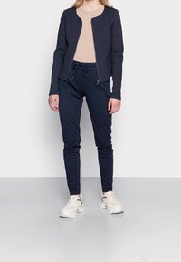 Chaqueta de fondo marino con cremallera y tela texturizada, parte superior beige y pantalones oscuros. Completada con zapatillas claras que presentan un patrón decorativo.