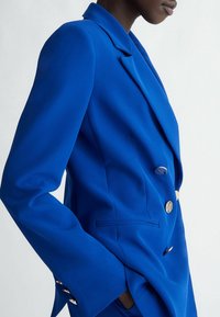Niebieska marynarka na miarę z klapami z wycięciami, dwurzędowym zapięciem, złotymi guzikami i usztywnionymi rękawami. Gładka tkanina i minimalistyczne kieszenie.
