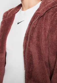 Bordowy płaszcz z sztucznego futra z kapturem i suwakiem, w zestawieniu z białym t-shirtem o prążkowanej teksturze, na którym znajduje się małe czarne logo Nike na piersi.