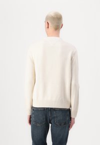 Pulover de culoare crem, cu tiv și manșete texturate, având un design posterior fără cusături și un material texturat. Asortat cu blugi de culoare albastru închis.