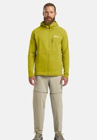 Haut à zip vert avec un tissu texturé et une poche frontale, associé à un pantalon beige clair avec des détails de couture et des zip sur les côtés.