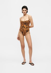 Γυναίκα που φοράει ένα ολόσωμο μαγιό με καφέ και μαύρες ρίγες τιγρέ και μαύρα σανδάλια τύπου slide, στέκεται με το ένα χέρι πίσω από την πλάτη της.