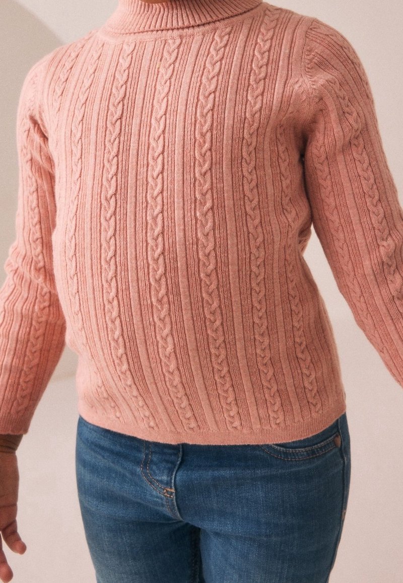 Enfant portant un pull en maille torsadée rose et un jean bleu, debout devant un fond uni.
