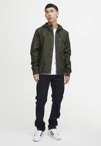 Hooded olive green jacket with zippered pocket, paired with a white t-shirt and dark jeans. Model wears white sneakers with black stripes.