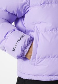 Doudoune isolée lavande avec poignets élastiques, dotée d'une poche latérale et du logo "Helly Hansen" en noir sur la manche.