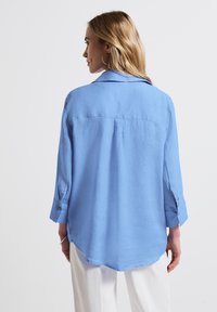 Camicia azzurro chiaro a maniche lunghe con piega sul retro e orlo arrotondato, caratterizzata da un colletto classico e dettagli con polsini abbottonati.