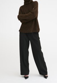 Pull à col roulé en maille marron, à coupe ample, associé à un pantalon noir taille haute à jambes larges et des escarpins noirs à bouts pointus.