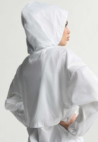 White lightweight jacket with a hood, featuring a cropped design and elastic cuffs. Smooth texture and minimalist style.