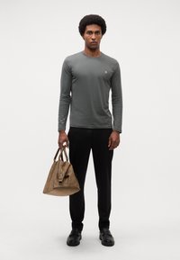 Maglietta grigia a maniche lunghe con scollo rotondo, abbinata a pantaloni neri. La modella tiene una borsa in pelle marrone. Le scarpe nere completano il look.