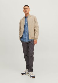Veste d'aviateur beige avec une poche zippée, associée à une chemise en denim bleu et un pantalon gris. Fini avec des sneakers blanches à accents bleus.