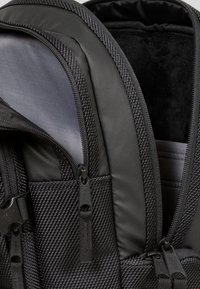 Gros plan d'un sac à dos noir avec plusieurs compartiments zippés et des détails en tissu texturé.