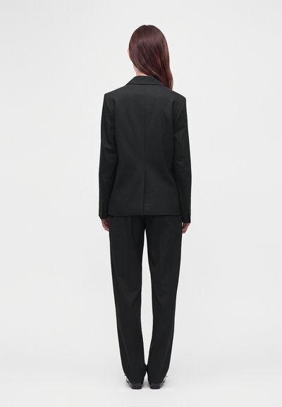 Femme aux longs cheveux bruns portant un costume noir ajusté vue de dos sur un fond clair uni.