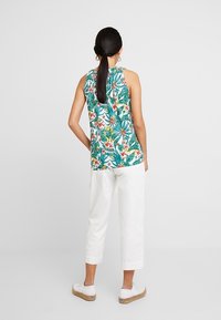 Vihreä ja valkoinen ilmanhihaa kukkapaita trooppisilla kuvioilla, yhdistettynä valkoisiin lyhyisiin housuihin ja vaaleanvärisiin kenkiin.