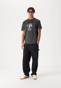 Grijze katoenen t-shirt met grafische print, gecombineerd met donkere, losse jeans. Het model draagt stijlvolle zonnebril en crèmewitte schoenen met rode strepen.