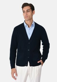 Cardigan blu navy a costine con scollo a V, cinque bottoni e due tasche frontali; indossato sopra una camicia azzurro chiaro, con un motivo lavorato a maglia texturizzato.