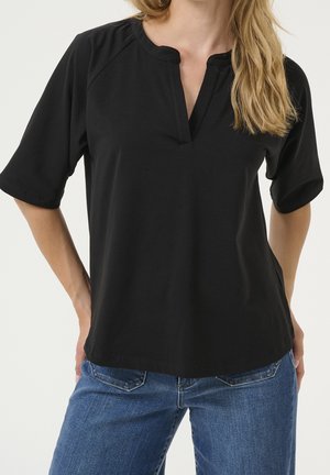 Frau trägt ein schwarzes kurzärmeliges Oberteil mit einem Schlitz im Ausschnitt, kombiniert mit blauen Jeans, steht mit den Händen hinter dem Rücken.