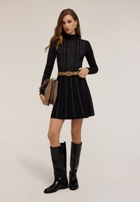 Rochie tricotată neagră cu prelungiri verticale din metal argintiu, mâneci lungi, guler înalt și fustă evazată. Curea maro la talie. Modelul poartă o geantă maro cu două compartimente. Cizme negre până la genunchi.
