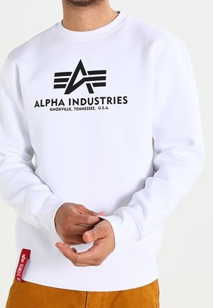 Sweat à capuche blanc en mélange de coton, avec un logo graphique noir et du texte, des poignets côtelés et une petite étiquette rouge sur le côté.