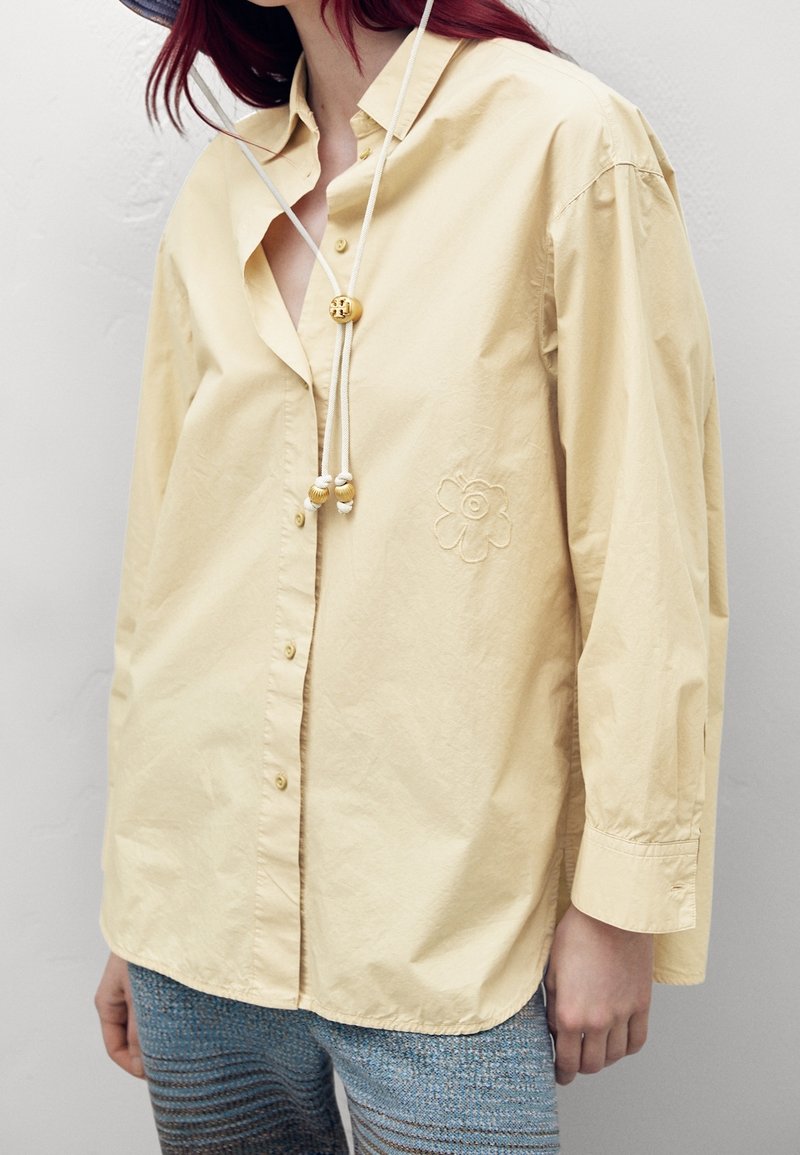 Femme portant une chemise ample jaune pâle boutonnée avec broderies florales et un pantalon bleu texturé.
