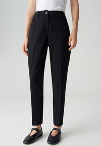 Personne portant un pantalon noir ajusté jusqu'à la cheville, un haut blanc sans manches, et des ballerines Mary Jane noires avec des brides à boucles, sur un fond uni.