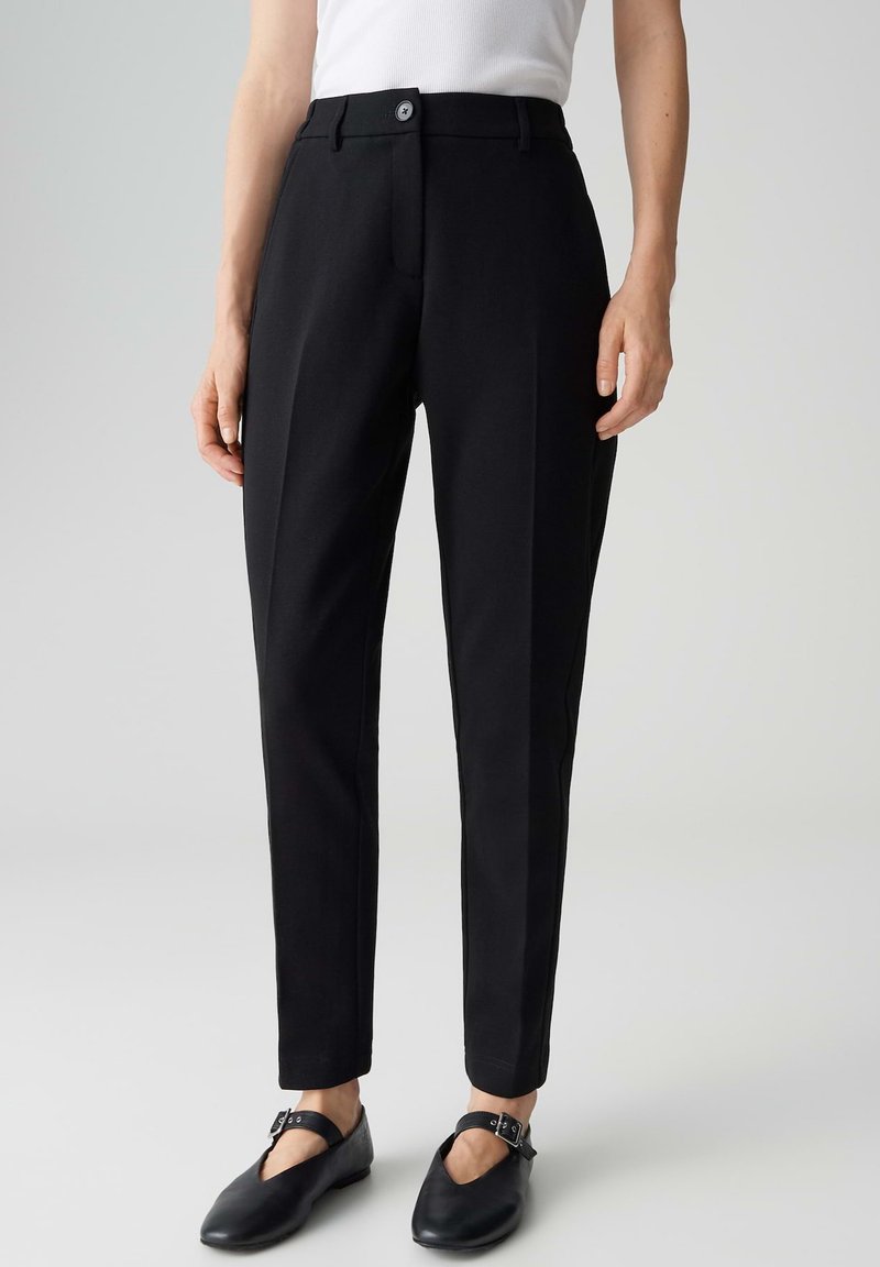 Personne portant un pantalon noir ajusté jusqu'à la cheville, un haut blanc sans manches, et des ballerines Mary Jane noires avec des brides à boucles, sur un fond uni.