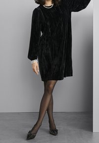 Femme portant une robe longue noire en velours avec un collier de perles et des bracelets, des collants noirs transparents et des chaussures noires pointues avec des nœuds.