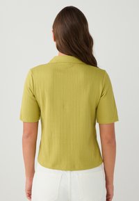 Kurzärmliges, kragenloses Hemd aus leicht grünem geripptem Stoff. Figurbetonendes Design mit strukturiertem Muster, sichtbaren Nähten und glatter Oberfläche.