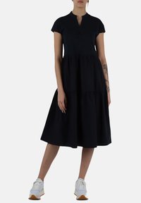 Abito nero a maniche corte con colletto e design a strati; realizzato in un tessuto fluido, abbinato a sneaker sportive bianche e grigie.