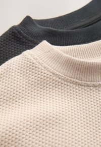 Tre teksturerede crewneck-strikketrøjer: en sort med et hævet mønster og to i beige med ribbet hals og glat tekstur.