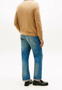 Homme portant un pull beige, un jean bleu et des mocassins noirs, debout, de dos, sur un fond blanc.