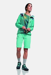 Leichte grüne Jacke mit Kapuze, kombiniert mit einem grün gemusterten Hemd und passenden Shorts. Schwarze und grüne Wanderschuhe runden den Look ab.