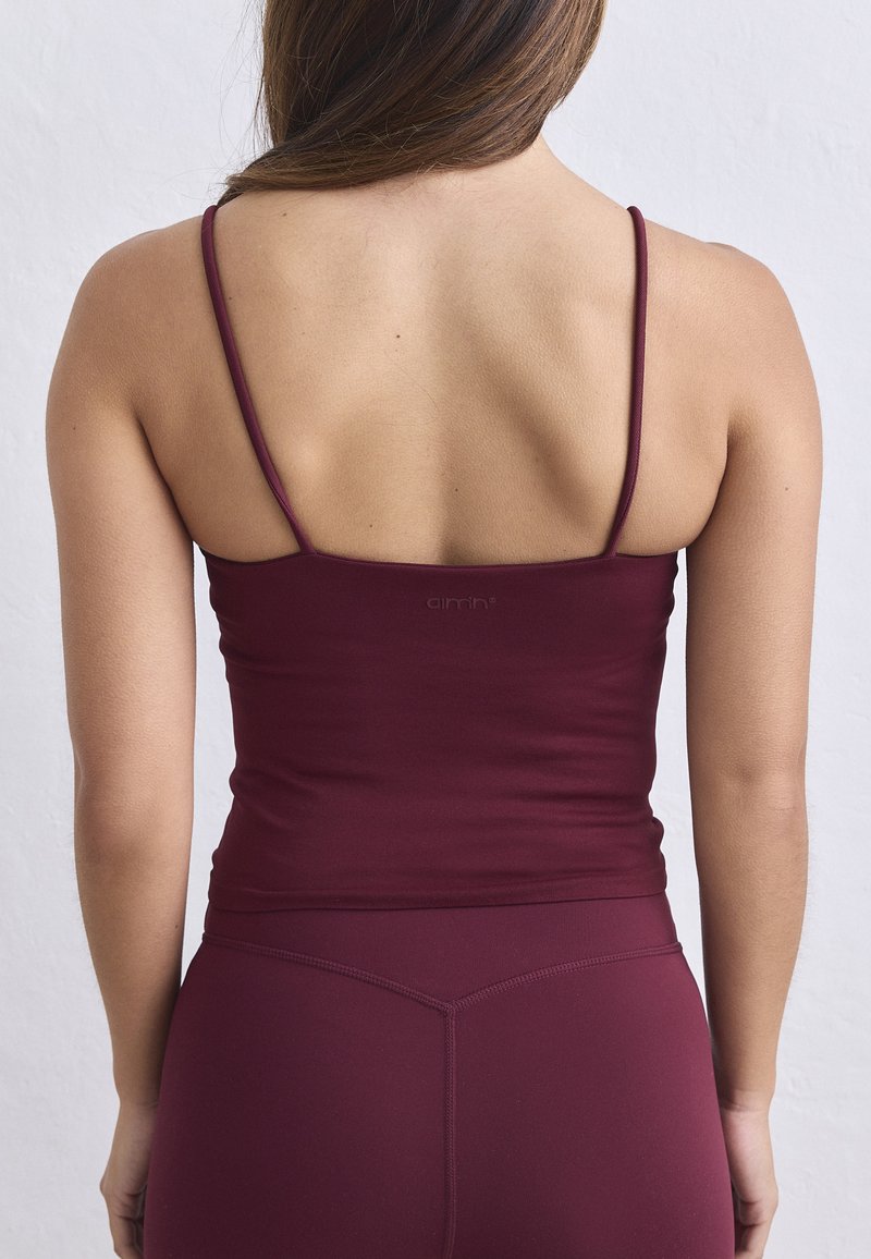 Femme portant un débardeur bordeaux ajusté avec des bretelles fines et des leggings assortis, vue de dos sur un fond uni.