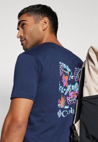 Mørkeblå t-skjorte laget av bomull med et blomstertrykkdesign som har rosa, blå og grønne blader på baksiden, med en logo nederst.