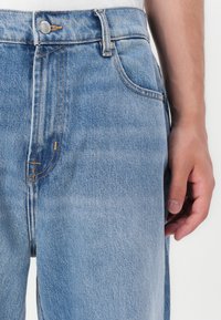 Jeans de mezclilla azul claro con un diseño clásico de cinco bolsillos, que presentan costuras naranjas y un cierre de botón en la cintura.