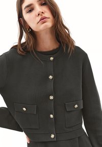 Dunkelgrüner, strukturierter Cardigan mit rundem Halsausschnitt, goldenen Knöpfen und zwei vorderen Taschen. Enganliegendes Design mit verkürzter Länge.