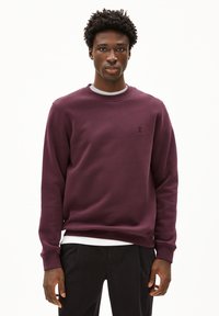 Felpa bordeaux con scollo rotondo, polsini a coste e un logo discreto. Tessuto dalla superficie liscia e vestibilità comoda, abbinata a pantaloni neri.