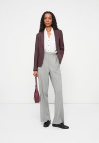 Blazer bordeaux sopra una camicia bianca con bottoni, abbinato a pantaloni grigi chiari con righe e scarpe nere. Il modello tiene in mano una piccola borsa bordeaux.